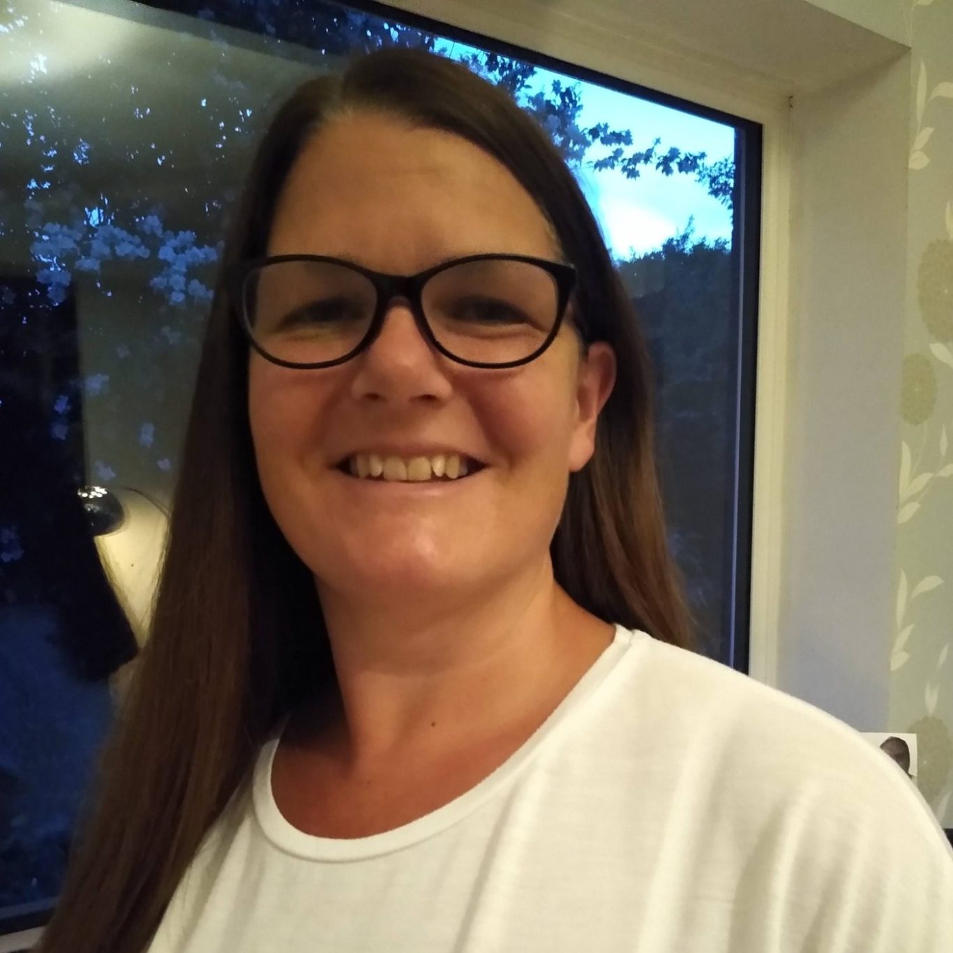 Smiling woman with long brown hair, wearing glasses, in a well-lit room.